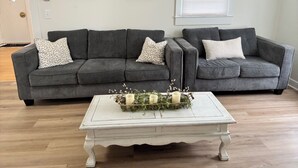 Ein Wohnzimmer mit einem grauen Ecksofa, zwei gerahmten floralen Kunstwerken, einem weißen Couchtisch mit Kerzen und Pflanzen sowie einer weißen Tür mit Klinke.