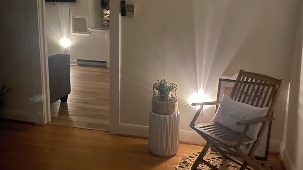 Ein gemütliches Zimmer mit Holzboden, einem Schaukelstuhl, einem kleinen runden Tisch mit Pflanze und einer Tür zu einem anderen Raum.
