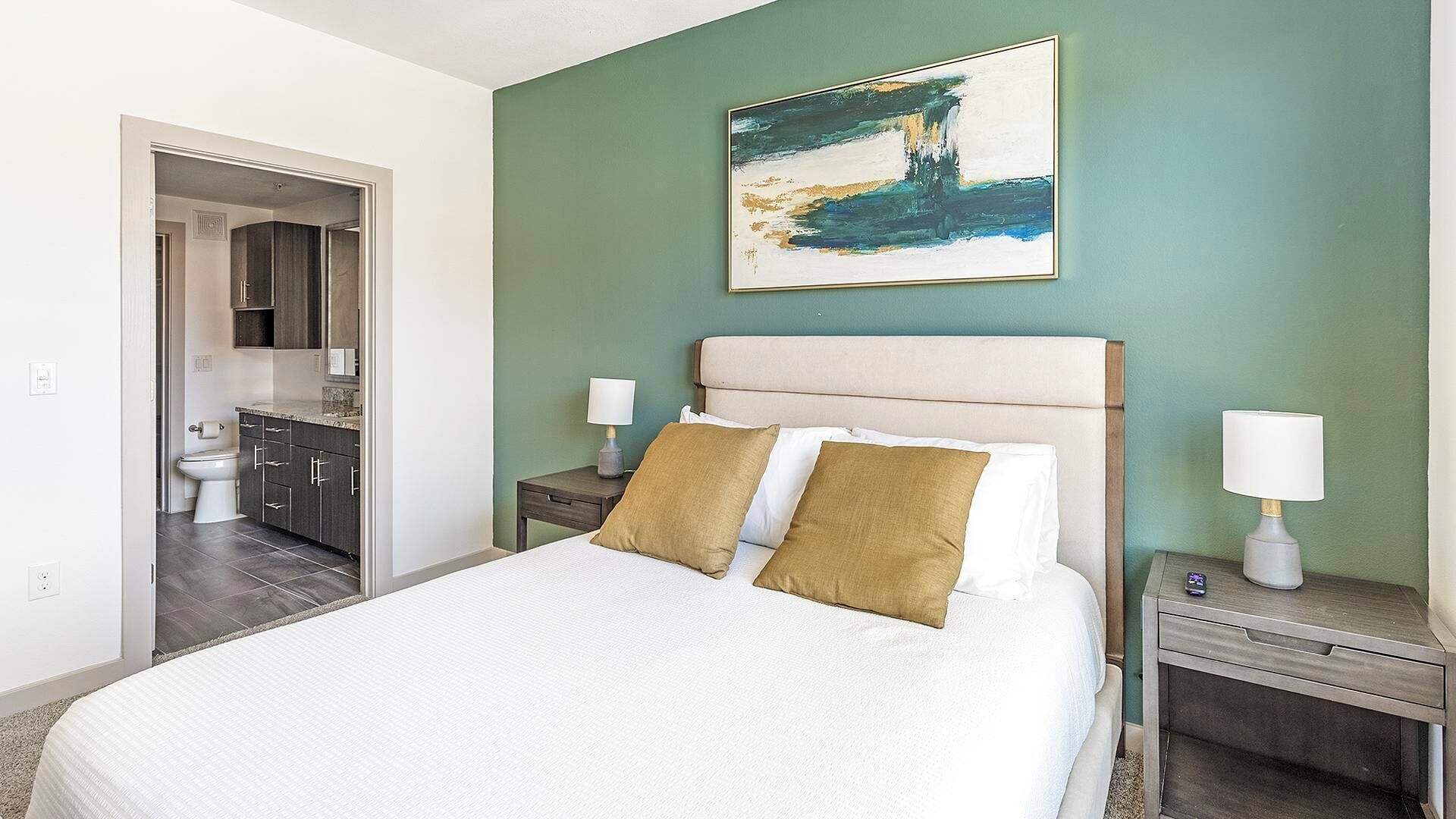 Habitación de hotel con cama, dos mesitas de noche con lámparas, un cuadro en la pared y un baño visible a través de una puerta abierta.