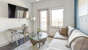 A modern living room with a glass coffee table, a sofa, a desk, and a TV mounted on the wall. - Landing | Amazing 1BD, Gym, Pool (Allen)