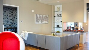 Una sala de estar moderna con un sofá gris, un sillón rojo y una mesa de centro de vidrio. Se observa una estantería con objetos decorativos y un cuadro en la pared.