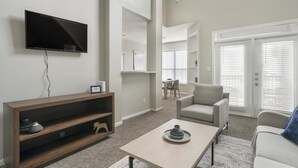 Un salon moderne avec un téléviseur à écran plat fixé au mur, un meuble de rangement en bois, un fauteuil gris, un canapé de couleur claire et une table basse agrémentée d’un objet décoratif.