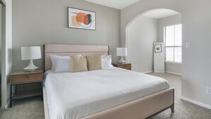 Une chambre avec un grand lit, deux tables de chevet avec des lampes, une œuvre d’art abstraite encadrée au-dessus du lit, et une porte en arc donnant sur une autre pièce.