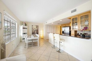 A modern kitchen with white cabinetry, a dining area with a white table and chairs, and a bar area with stools. - Beachfront Condo w/ Pool & Balcony Sunset Views (Clearwater)