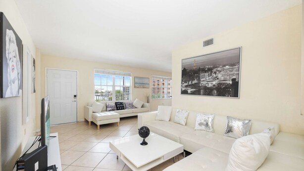 A modern living room with a white sofa, a coffee table, and a flat-screen TV. There are two windows with a view of buildings outside. - Beachfront Condo w/ Pool & Balcony Sunset Views (Clearwater)