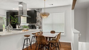 A modern kitchen with dark cabinets, a marble backsplash, a central island with stools, and a dining table with chairs. - Gothic Girl Stylish Upstairs Nook Near Shops & A&M (Bryan)