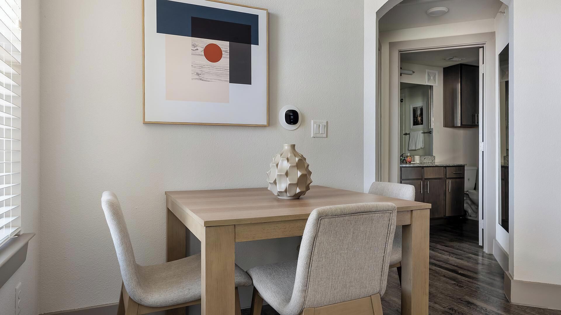 A dining area with a wooden table and two chairs, a vase on the table, and a framed artwork on the wall.
