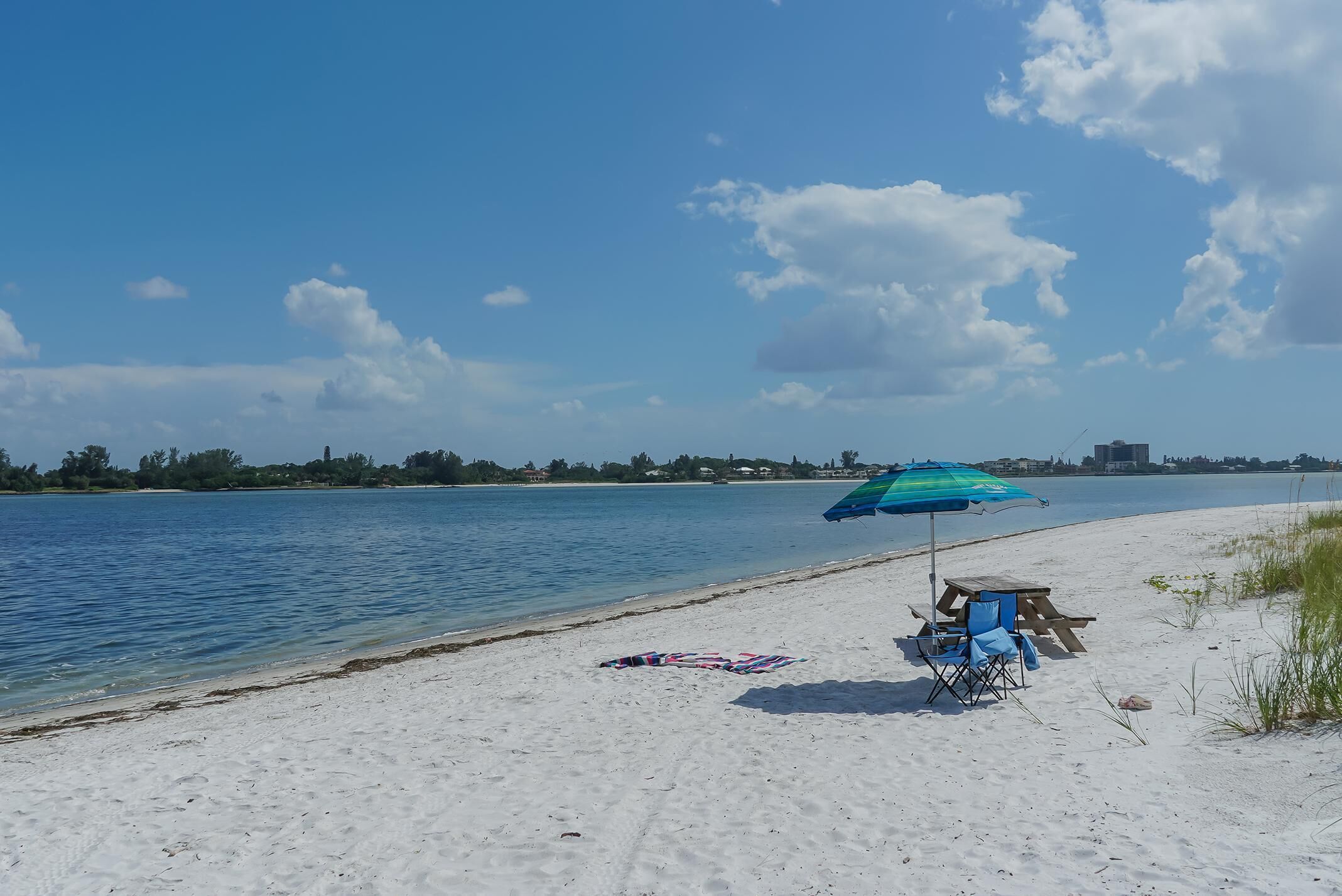 A beach with a clear sky, a wooden bench, a blue umbrella, and a beach chair.