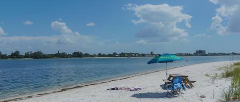A beach with a clear sky, a wooden bench, a blue umbrella, and a beach chair.