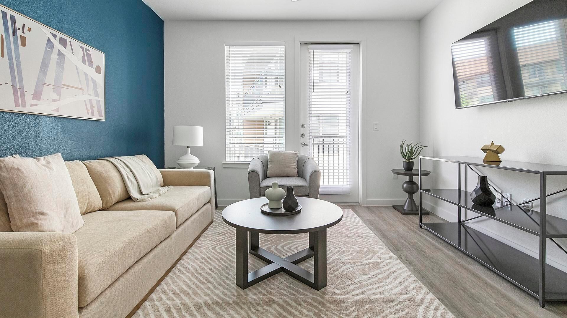 Un salon moderne avec un canapé beige, une table basse ronde et un fauteuil gris. Il y a deux fenêtres équipées de stores, un téléviseur fixé au mur et une petite table d’appoint avec une lampe.