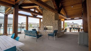 An outdoor patio with stone fireplace, blue chairs, and a pool in the background. - Landing | Modern 3BD, Gym, Pool (Waller)