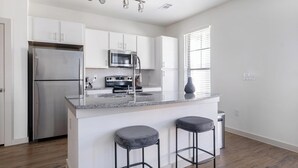 A modern kitchen with white cabinets, stainless steel appliances, a granite countertop, and two bar stools. - Landing | Modern 3BD, Gym, Pool (Waller)