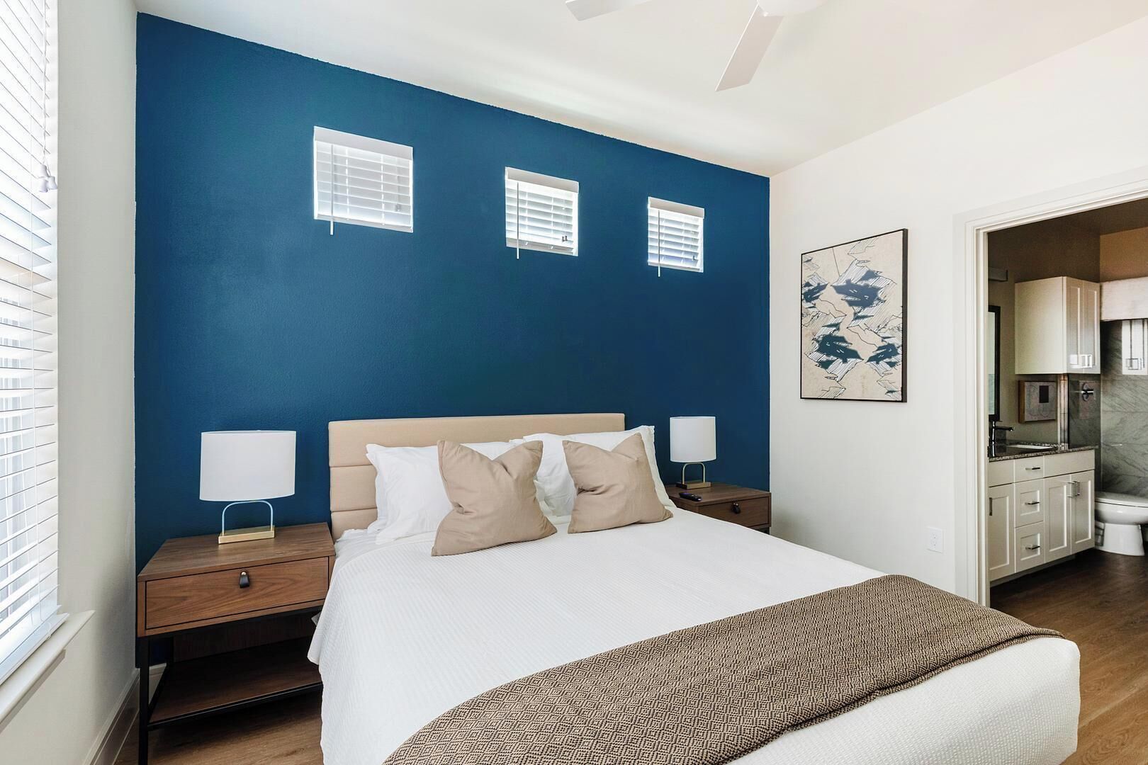 A bedroom with a large bed, two bedside tables, and a nightstand. There are three windows with blinds, a painting on the wall, and a bathroom visible through an open door.