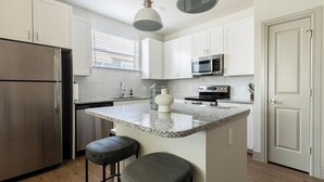 Una cocina moderna con armarios blancos, un refrigerador de acero inoxidable, un microondas y una isla de encimera de granito con dos taburetes de bar.