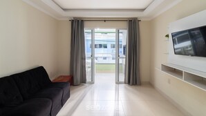 Un salon avec un canapé noir, un téléviseur à écran plat fixé au mur et une porte-fenêtre coulissante donnant sur un balcon extérieur.
