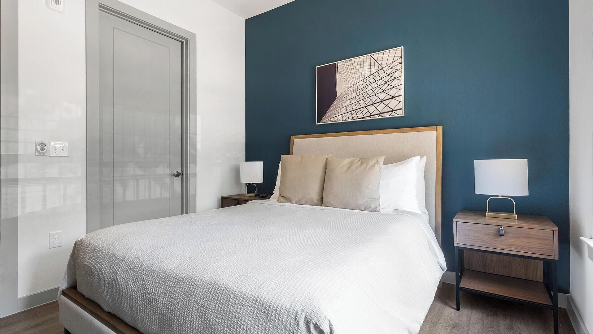 A bedroom with a bed, two bedside tables, a nightstand, a wall-mounted artwork, and a door.