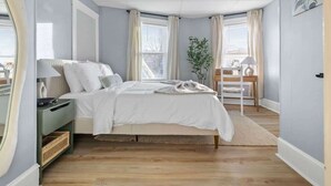 Quarto com cama, mesas de cabeceira, um criado-mudo, janela com cortinas, uma planta e uma vista.