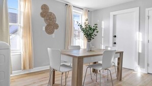 Uma sala de jantar com mesa de madeira e cadeiras brancas, uma planta em vaso e uma parede com arte decorativa.