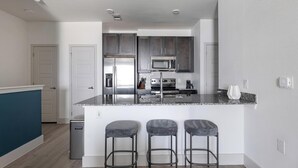 A modern kitchen with stainless steel appliances, a granite countertop, and three bar stools. - Landing | Stunning 1BD, Gym, Pool (Austin)
