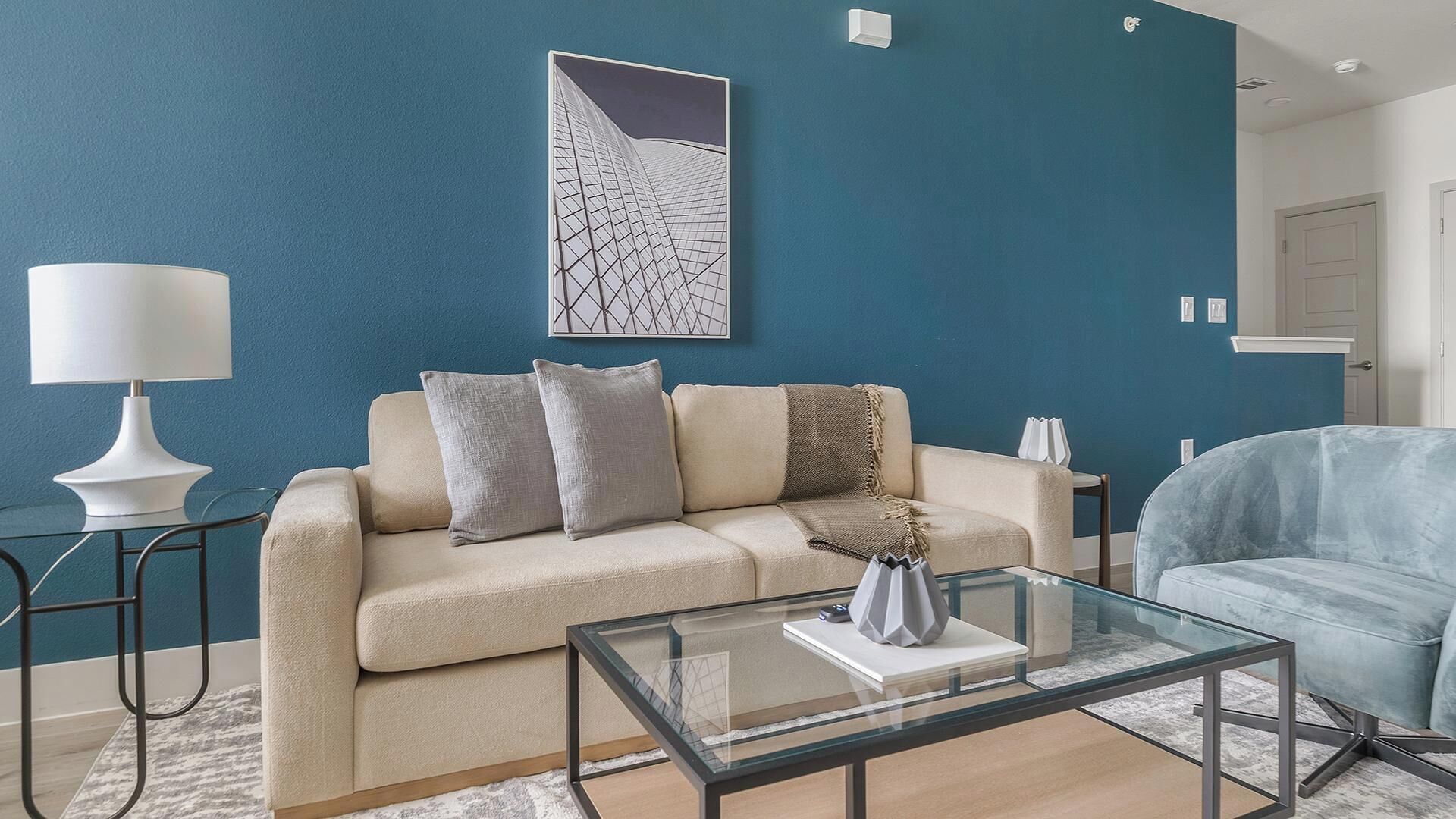 A modern living room with a blue accent wall, a beige sofa, a glass coffee table, and a blue armchair.