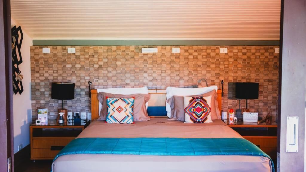 Quarto de hotel com parede estampada, cama com manta azul, duas mesas de cabeceira com abajures e almofadas decorativas.