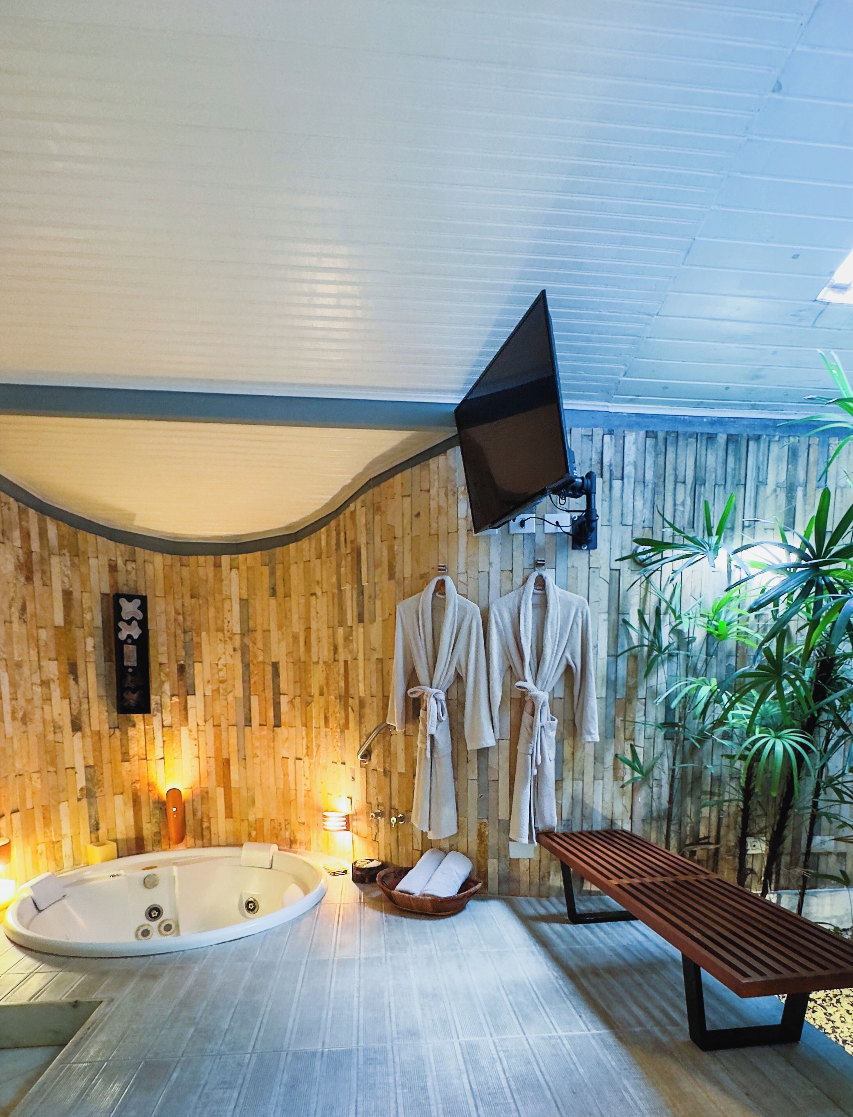 A spa room with a wooden wall, a large bathtub, two white robes, a bench, and a TV mounted on the wall.