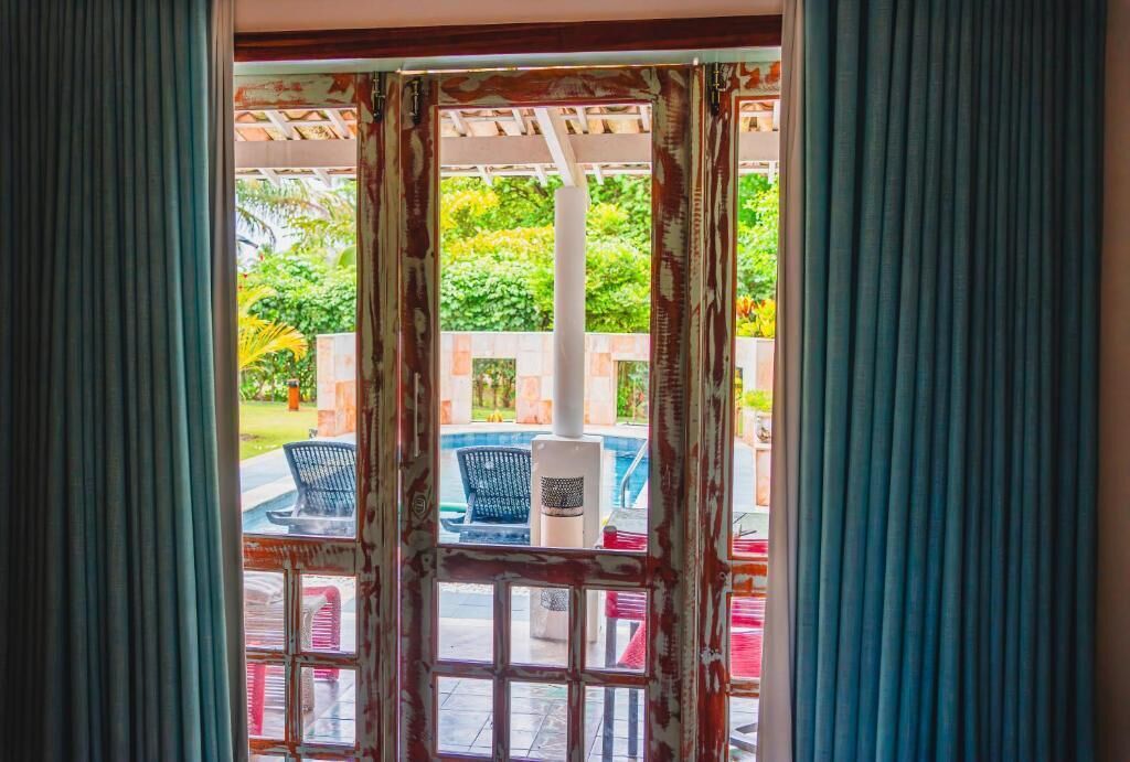 Une vue à travers une fenêtre avec un cadre en bois et des rideaux, donnant sur un espace extérieur avec une piscine, des chaises et une table.