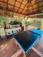 Habitación con techo de paja, una mesa de masajes, un estante con adornos y abundante vegetación afuera.