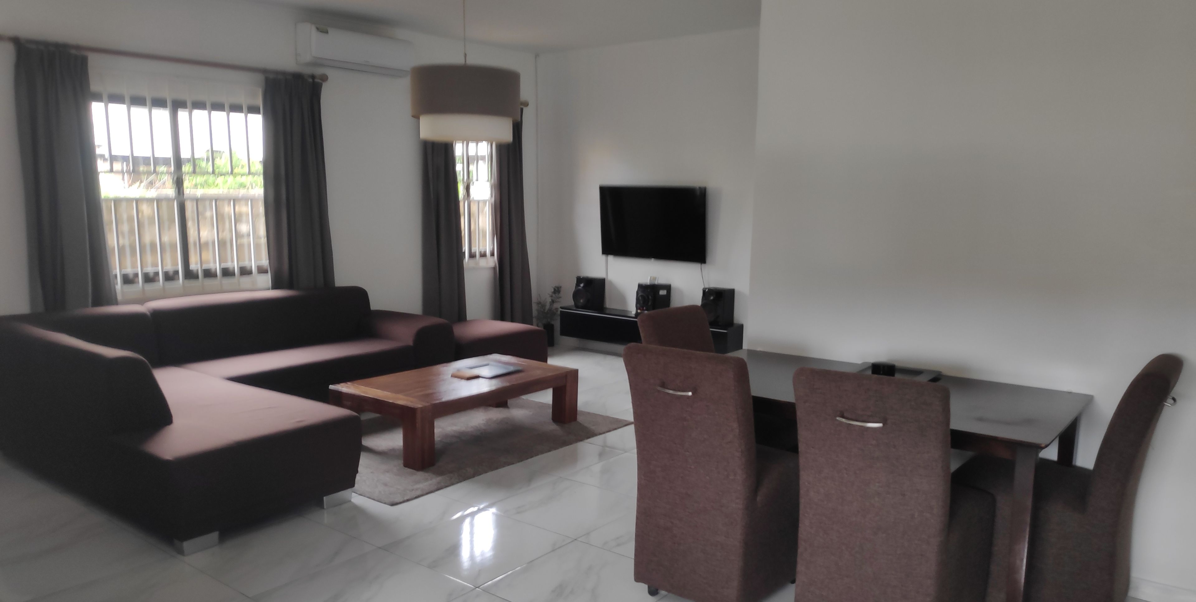 Una sala de estar moderna con un sofá modular, una mesa de centro y dos sillones. Hay un televisor de pantalla plana montado en la pared, un ventanal con cortinas y un equipo de aire acondicionado.