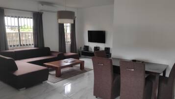 Una sala de estar moderna con un sofá modular, una mesa de centro y dos sillones. Hay un televisor de pantalla plana montado en la pared, un ventanal con cortinas y un equipo de aire acondicionado.
