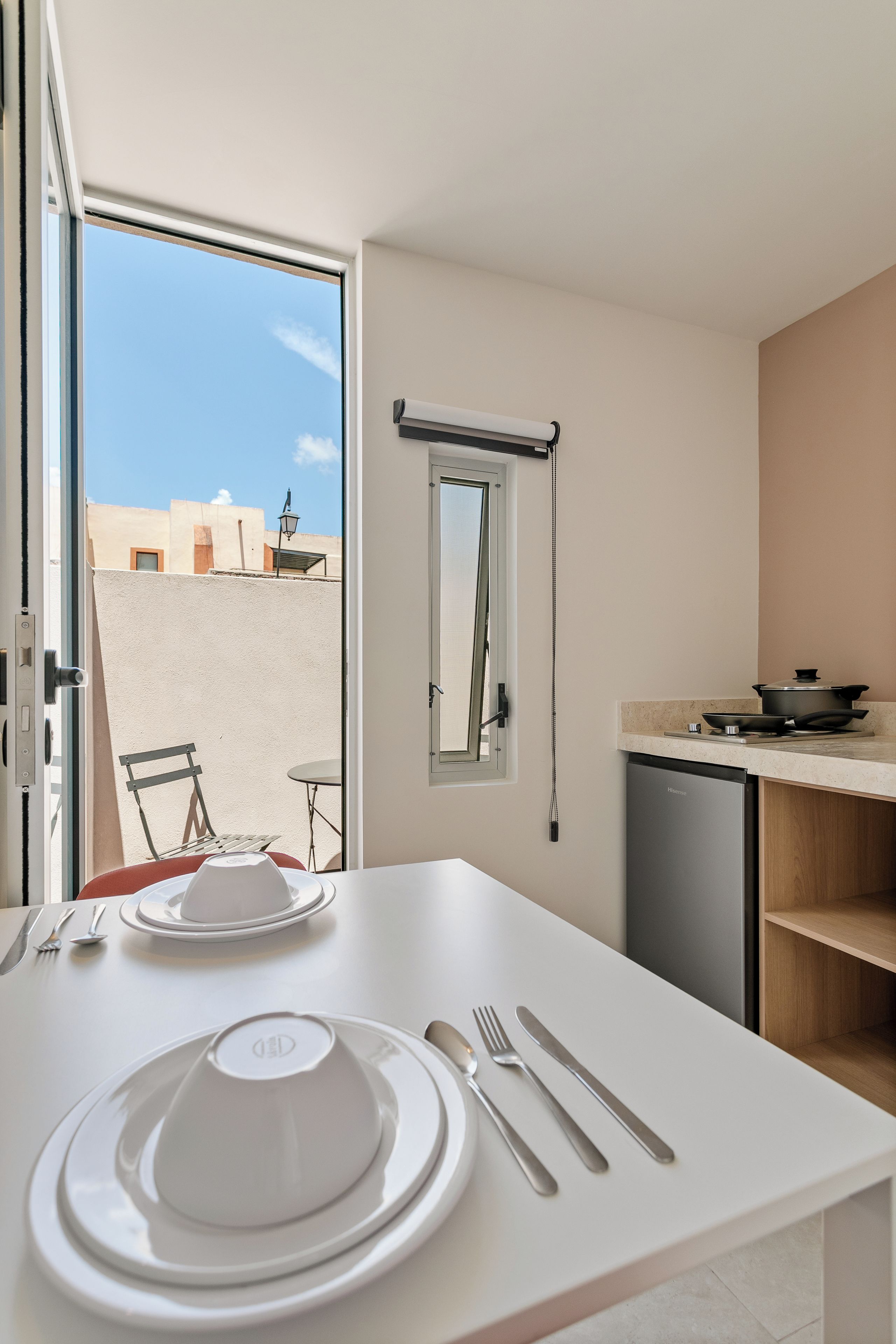 A compact kitchen with a dining area, a small table set with plates, cutlery, and a cup. A window with a view of buildings is visible, and a ceiling fan is mounted on the wall.