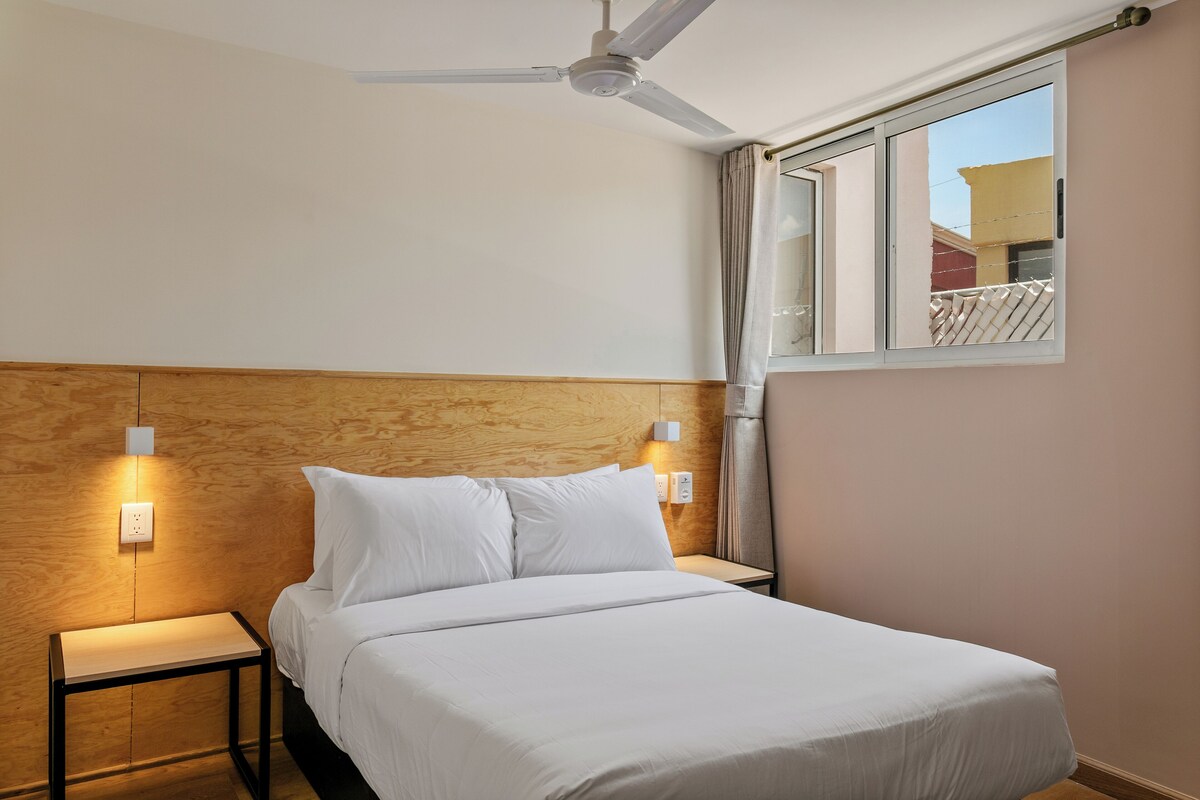 Una cama bien hecha con sábanas blancas, cabecera de madera, mesita de noche con lámpara y ventilador de techo.