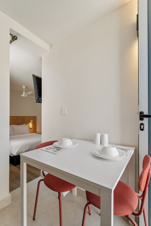 Quarto de hotel com uma mesa branca posta para dois, cadeiras vermelhas, cama com cabeceira de madeira e uma televisão na parede.