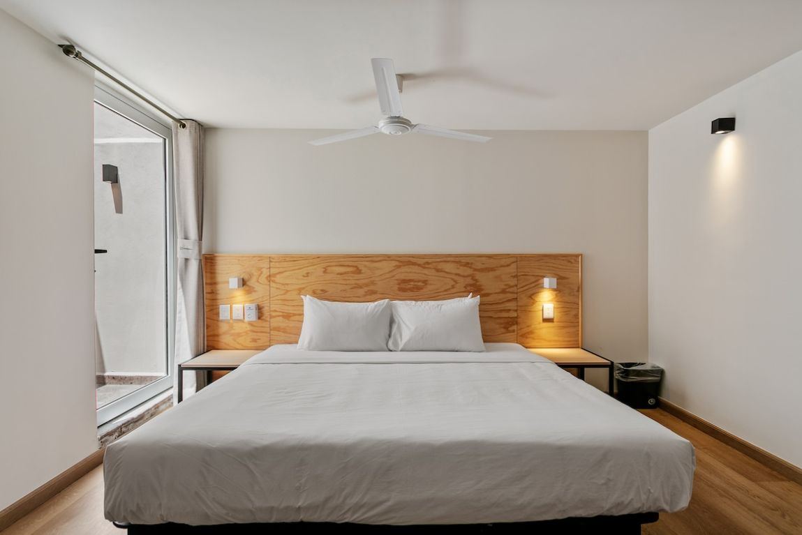 A hotel room with a bed, a ceiling fan, bedside lamps, and a window.