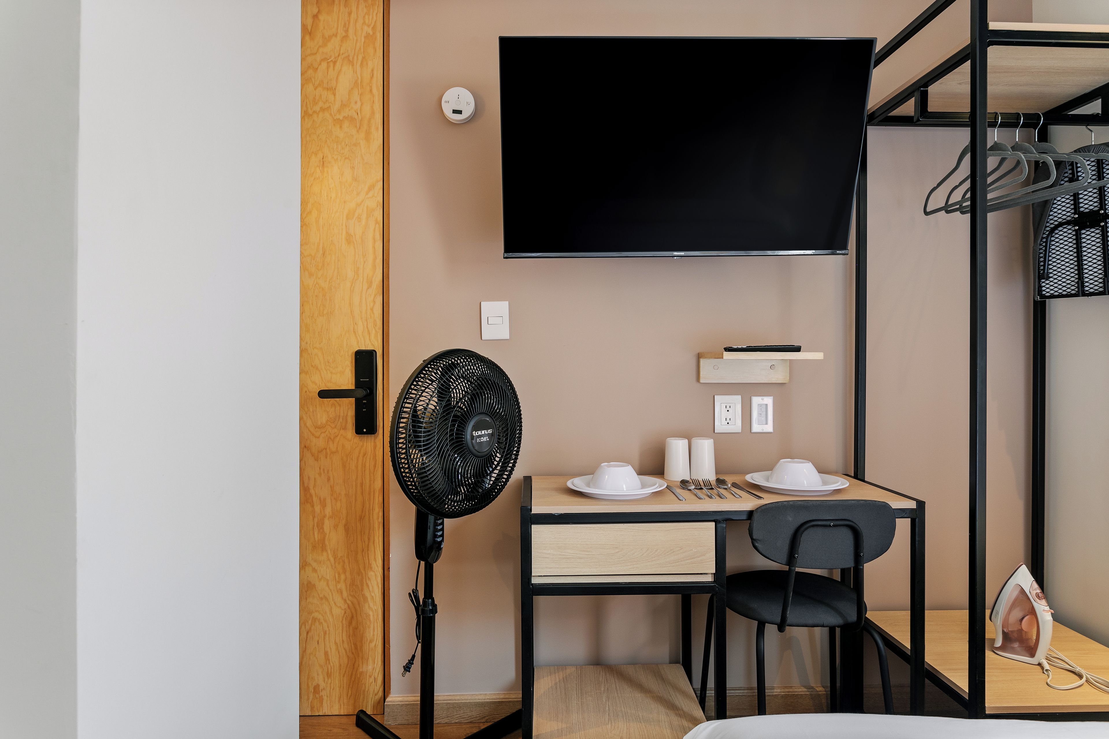 A compact hotel room with a bed, a desk with a chair, a television mounted on the wall, a standing fan, and a small ironing station.