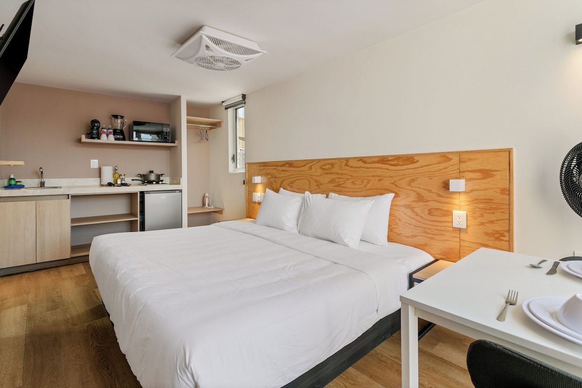 A hotel room with a bed, a desk with a chair, a small kitchenette, and a window.
