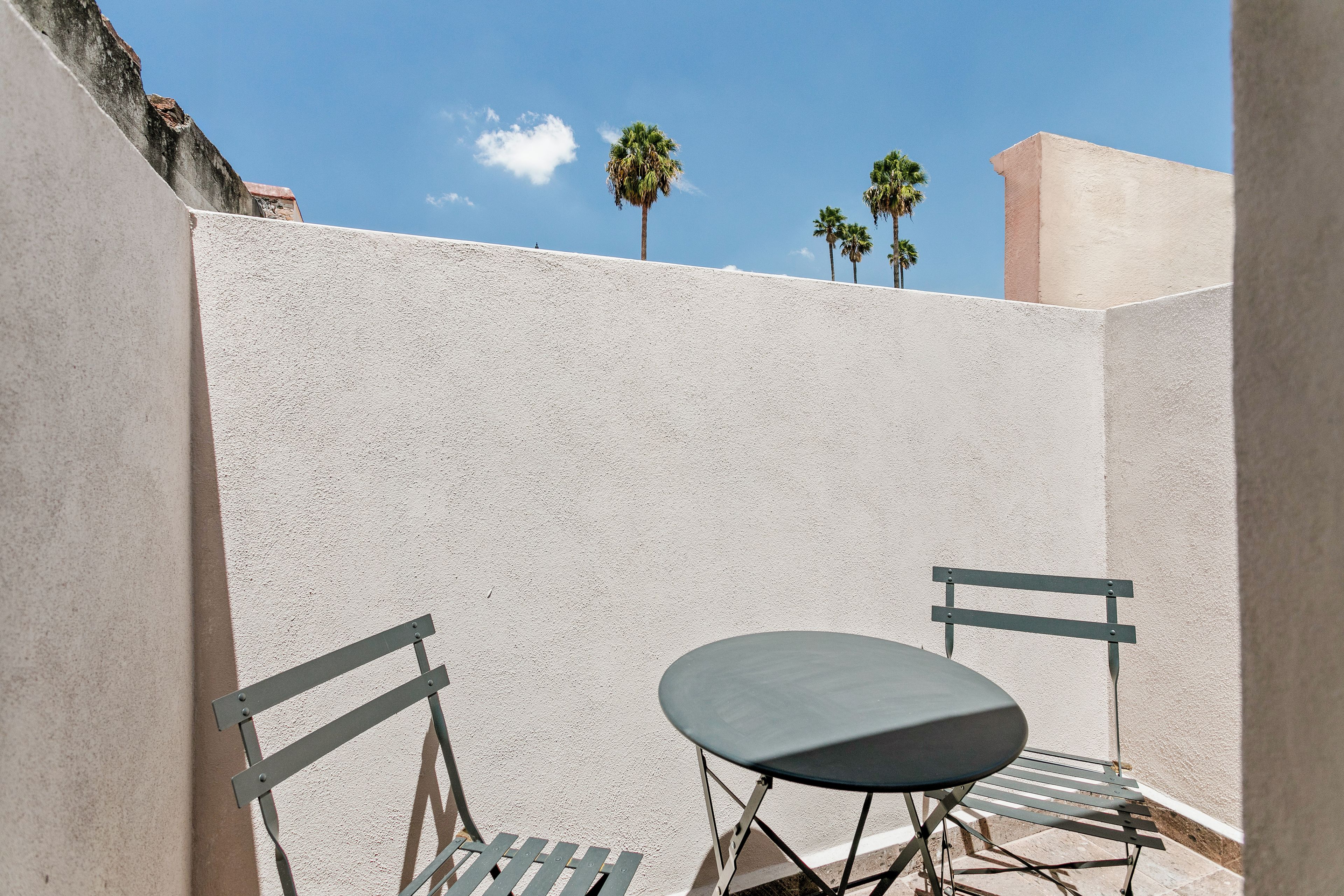 A small outdoor seating area with a table and two chairs, surrounded by a textured wall and palm trees in the background.