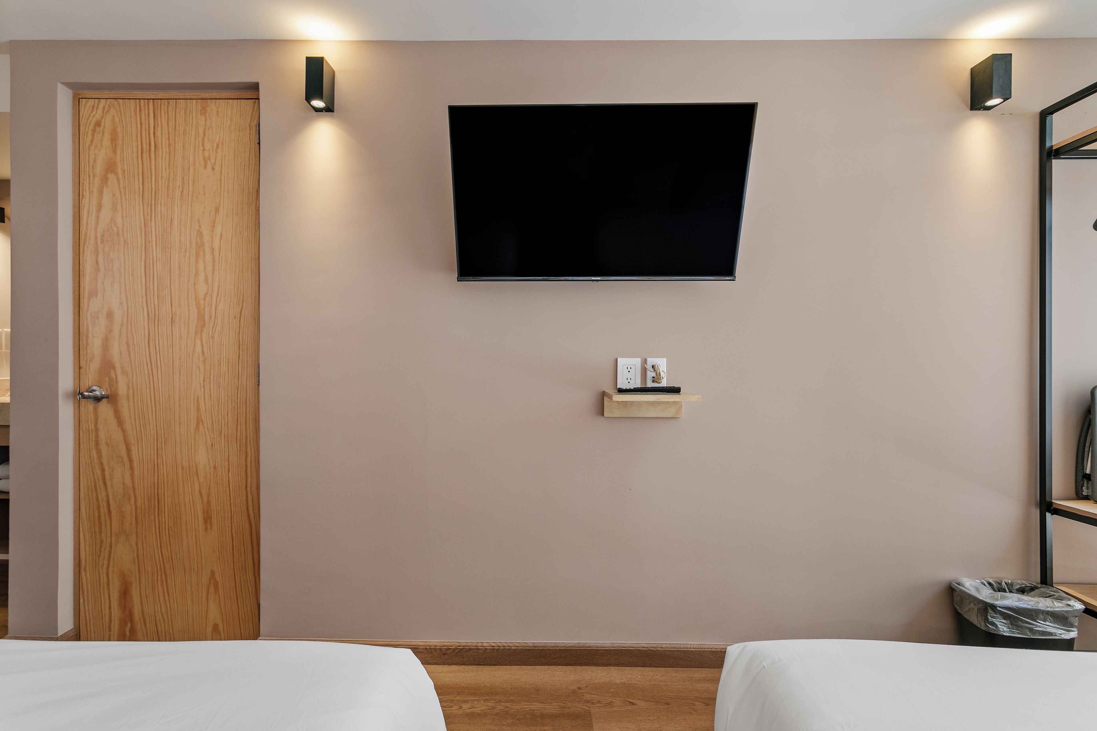 Habitación de hotel con puerta de madera, un televisor de pantalla plana montado en la pared, un estante pequeño con toma de corriente y un cubo de basura.