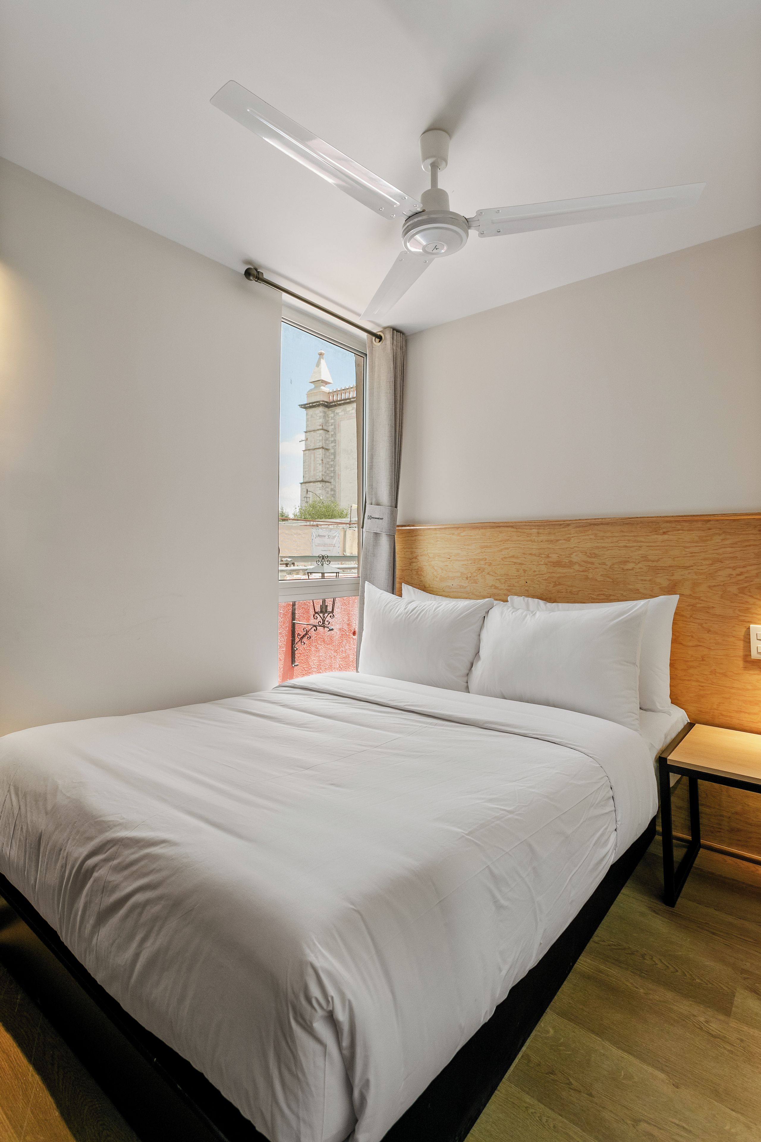 A hotel room with a bed, a nightstand, a ceiling fan, and a window with a view of a building.