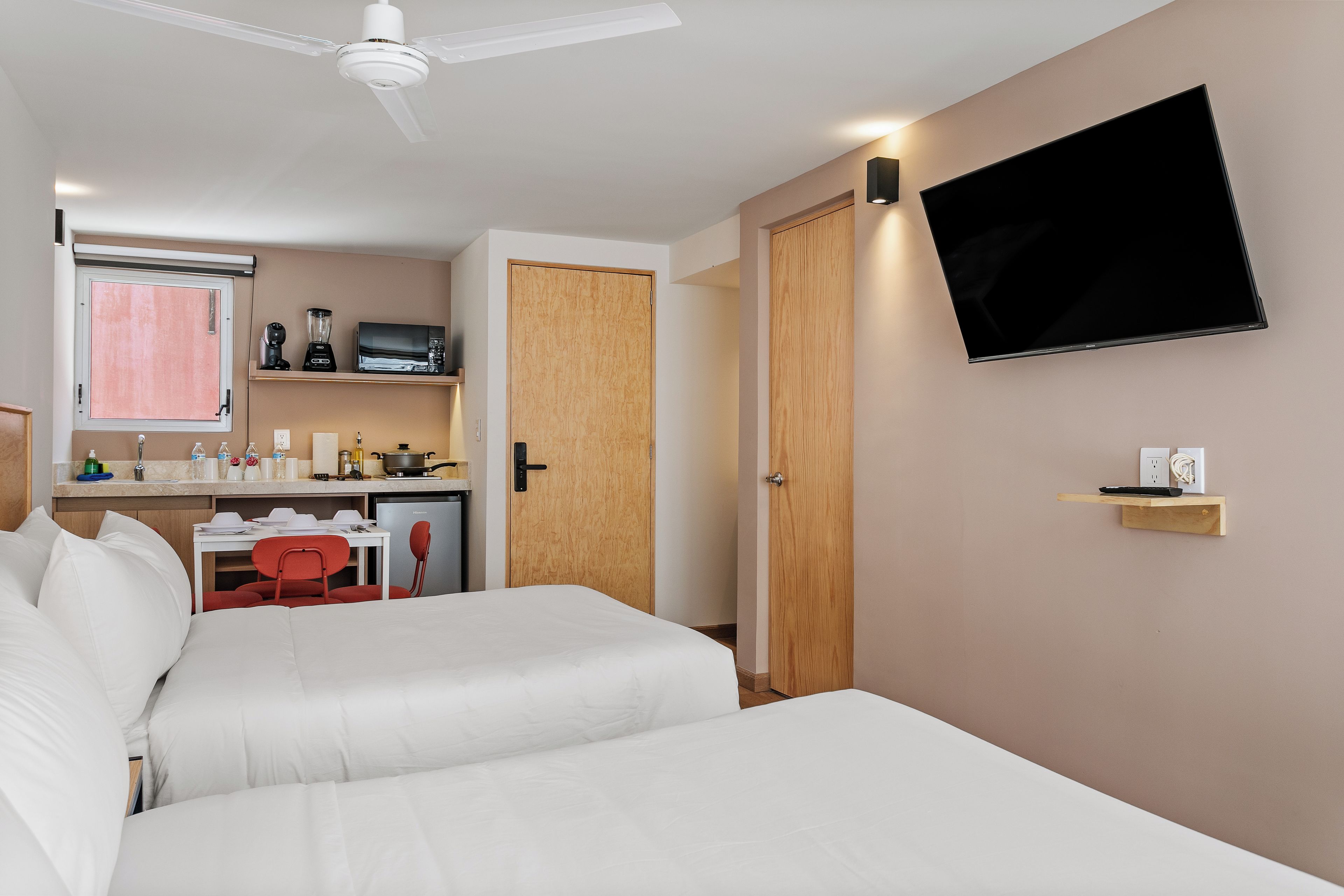 Una habitación de hotel con cama, televisión montada en la pared, kitchenette y ventilador de techo.