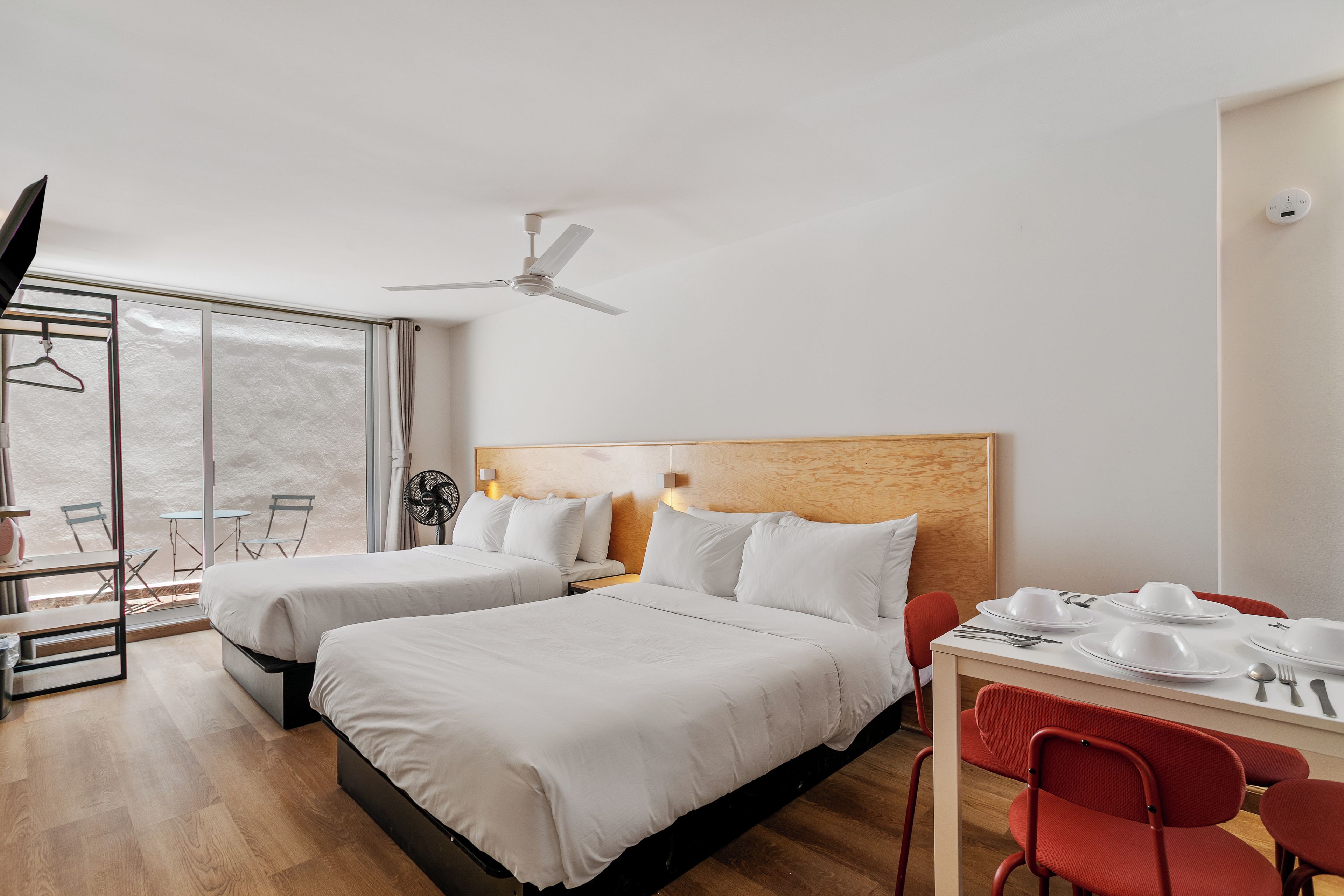 A hotel room with two beds, a dining table with chairs, a ceiling fan, and a large window.