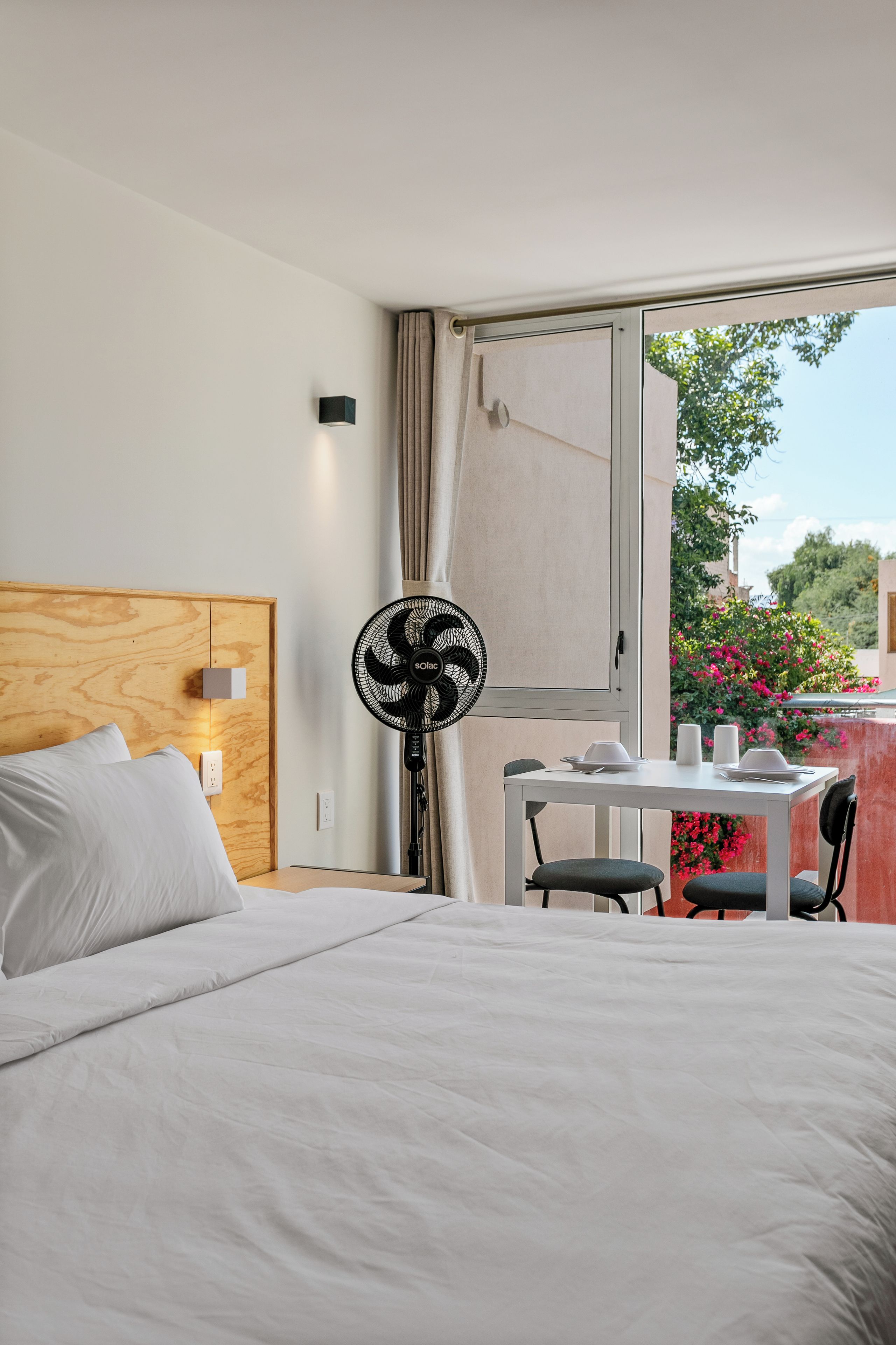 A hotel room with a bed, a table with chairs, a standing fan, and a window with curtains.