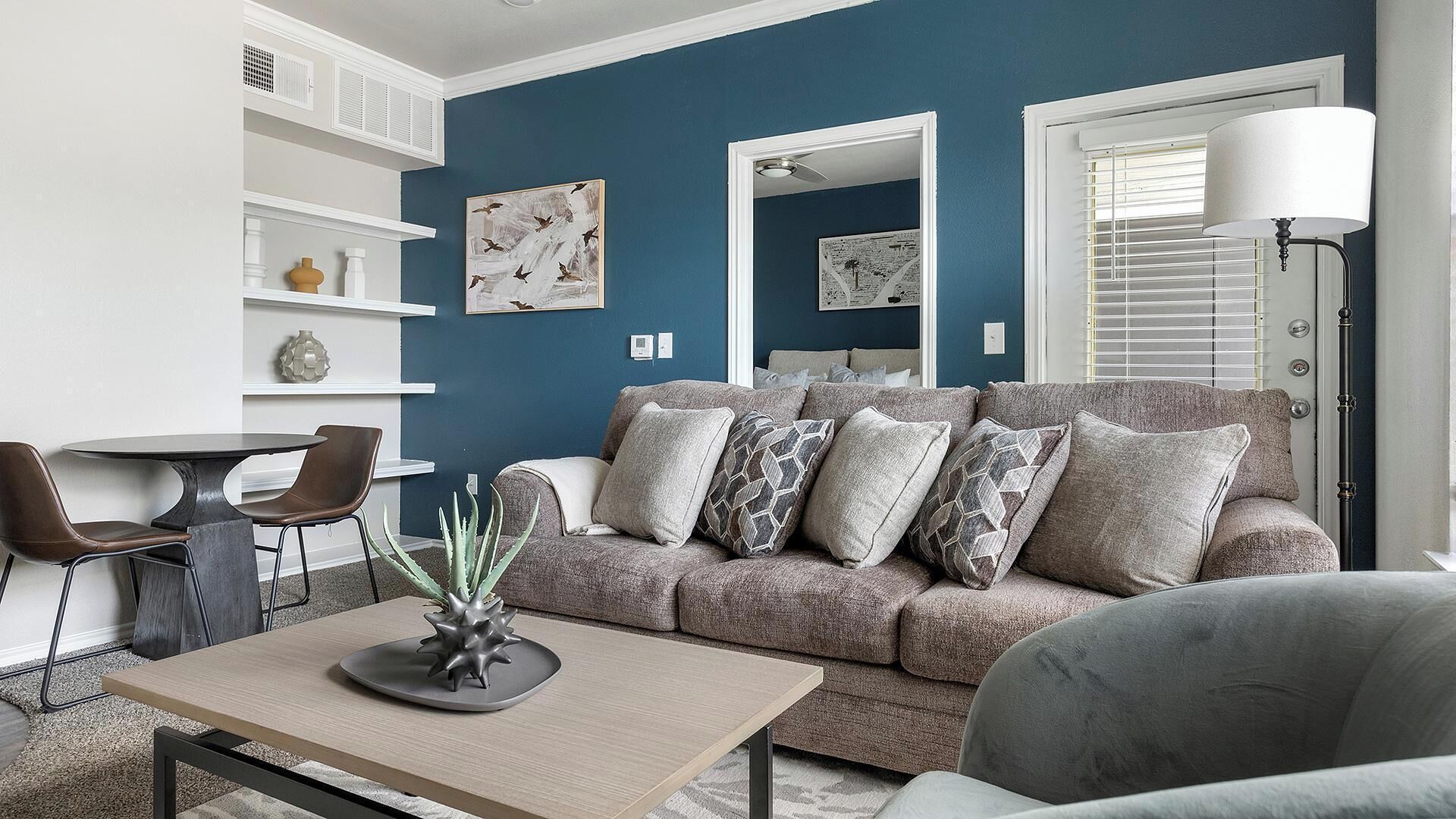A living room with a blue wall, a grey sofa with patterned cushions, a round coffee table, and a dining area with a white table and two chairs.