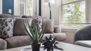 Ein Wohnzimmer mit grauem Sofa, gemusterten Kissen, einem Topf mit Aloe Vera und einer modernen Stehlampe.