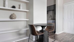 A dining area with a round table and two chairs, a white wall with shelves, and a kitchen in the background.