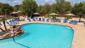 A swimming pool with blue lounge chairs and umbrellas, surrounded by a wooden fence and trees.