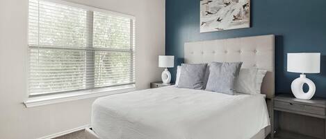 A bedroom with a large bed, two bedside tables with lamps, a window with blinds, and a wall-mounted artwork.