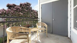 A balcony with wicker furniture, a view of pink flowering trees, and a grey door. - Landing | Modern 1BD, Gym, Clubhouse (San Antonio)