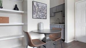 A dining area with a round table and two chairs, a framed cityscape picture on the wall, and a kitchen visible through an open door. - Landing | Modern 1BD, Gym, Clubhouse (San Antonio)