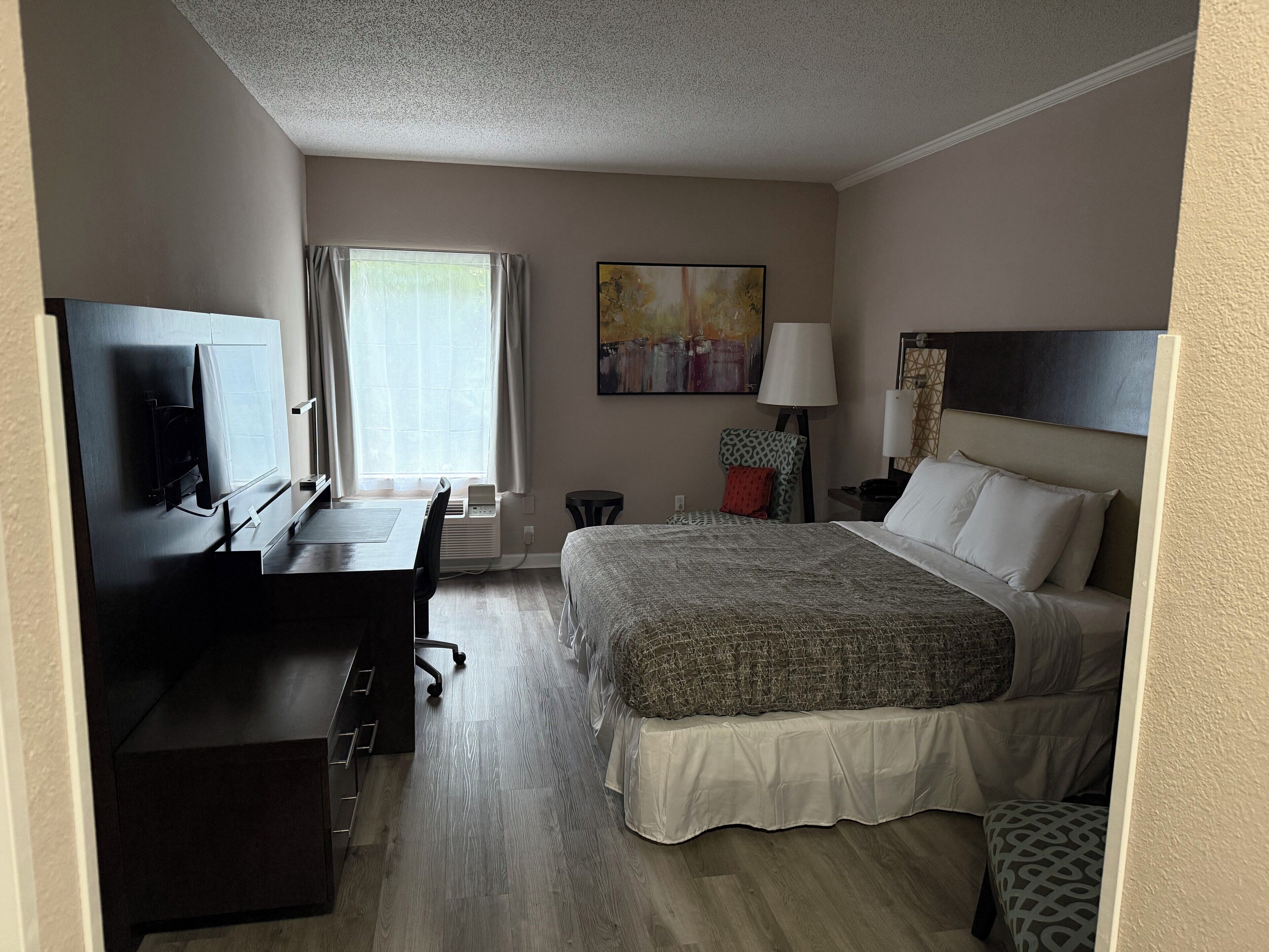 A hotel room with a bed, desk, chair, TV, and artwork on the wall.